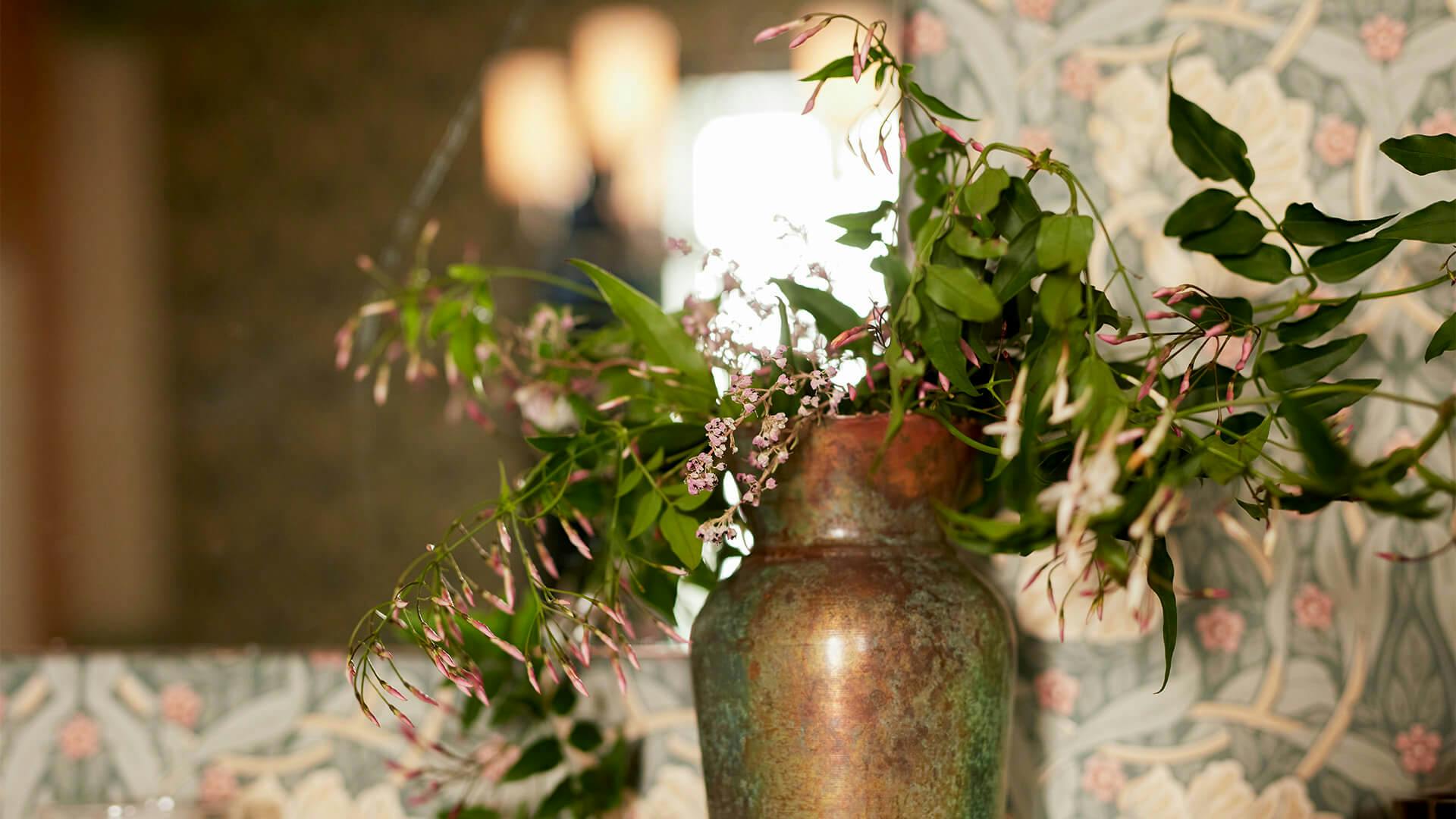 Vase with flowers in front of wall with decorative wallpaper and large mirror.